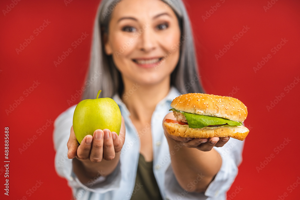 Close-up portrait of mature senior asian woman holding burger and green apple. Standing isolated over red background. Diet concept. Close-up portrait of mature senior asian woman holding burger and green apple. Standing isolated over red background. Diet concept.