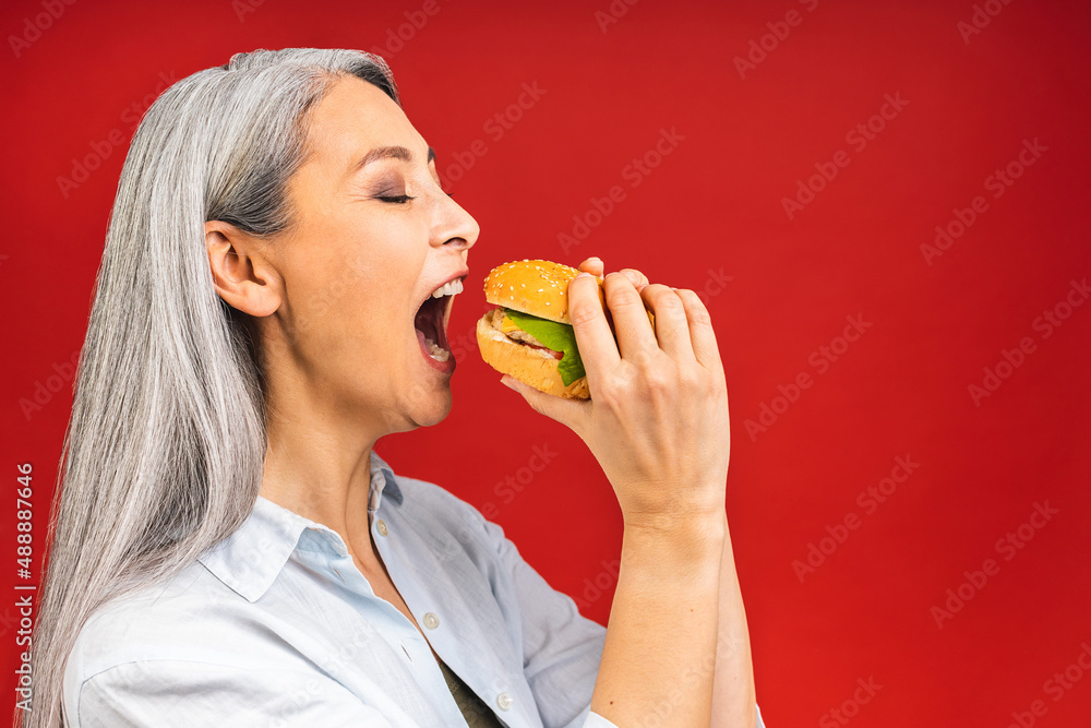 Mature senior woman eating burger with satisfaction. Grandmother enjoys tasty hamburger takeaway, delicious bite of burger, order fastfood delivery while hungry, standing isolated over red background. Mature senior woman eating burger with satisfaction. Grandmother enjoys tasty hamburger takeaway, delicious bite of burger, order fastfood delivery while hungry, standing isolated over red background.