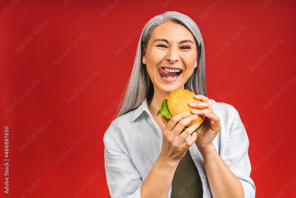 Mature senior asian woman eating burger with satisfaction. Grandmother enjoys tasty hamburger takeaway, order fastfood delivery while hungry, standing isolated over red background. Mature senior asian woman eating burger with satisfaction. Grandmother enjoys tasty hamburger takeaway, order fastfood delivery while hungry, standing isolated over red background.