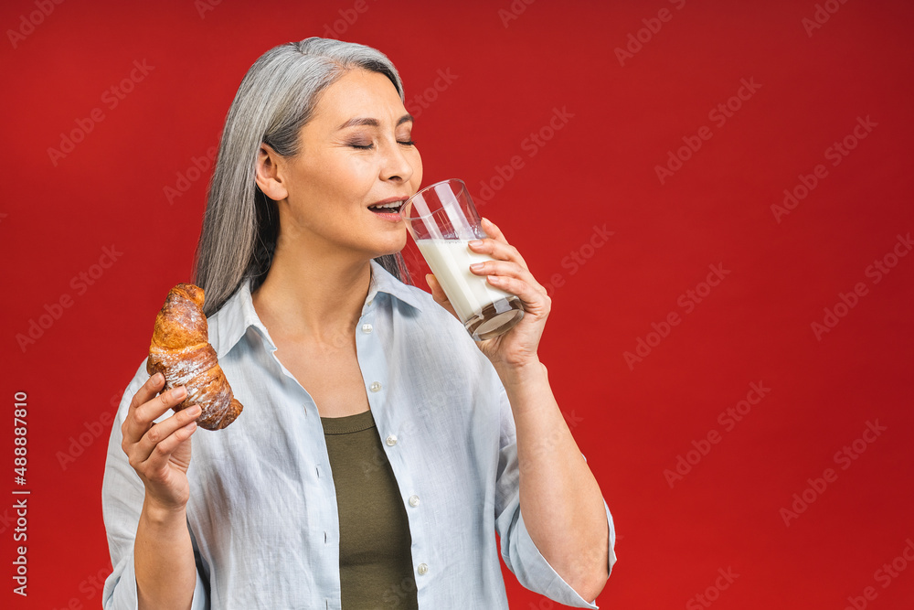 Asian senior mature aged woman drinking milk for calcium food and healthy lifestyle concept isolated on red background. Lady eating croissant.