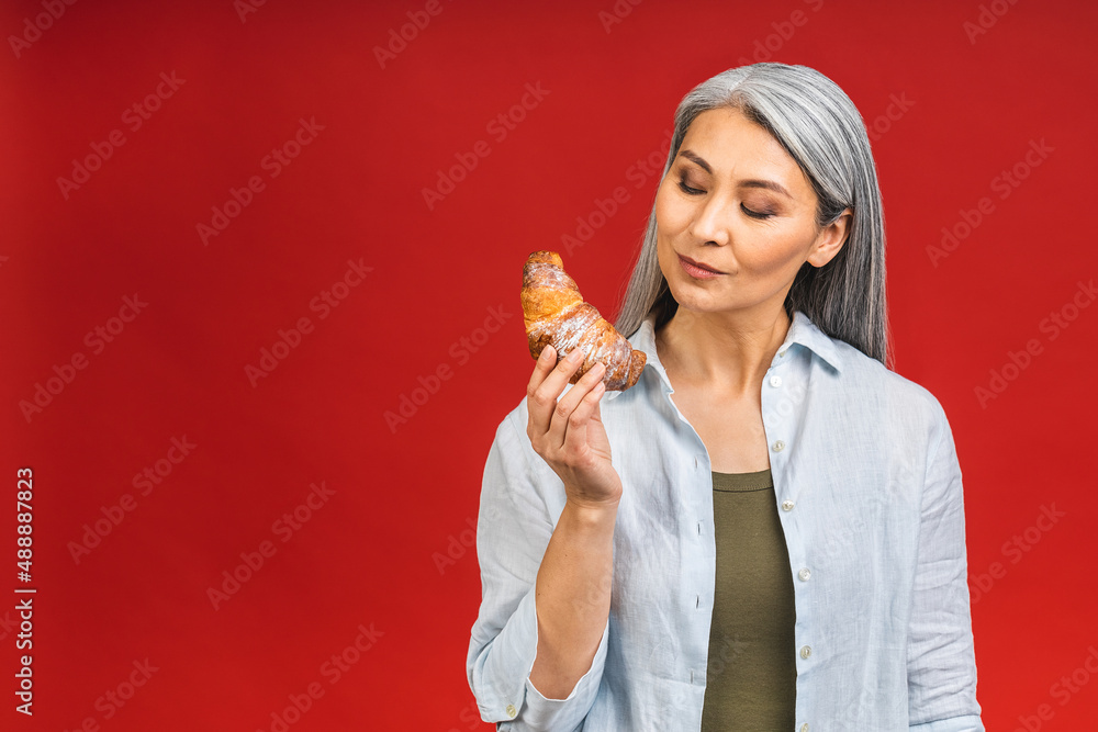 Portrait of a happy asian senior mature aged woman eating croissant isolated over red background. French breakfast concept. Portrait of a happy asian senior mature aged woman eating croissant isolated over red background. French breakfast concept.