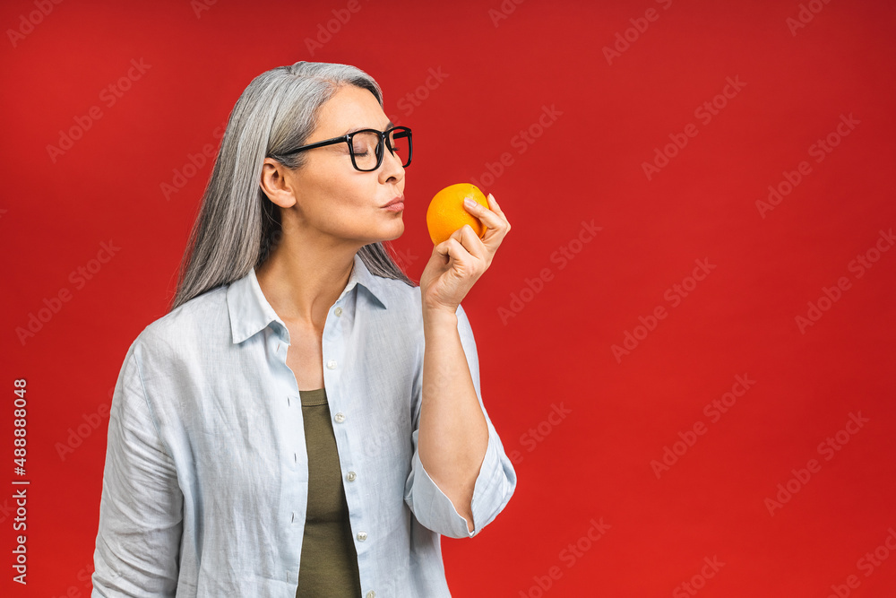 Vegan or vegetarian concept. Portrait of a beautiful elderly asian mature aged woman holding an orange fruit, smiling, isolated over red background. Vegan or vegetarian concept. Portrait of a beautiful elderly asian mature aged woman holding an orange fruit, smiling, isolated over red background.
