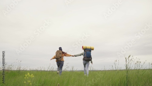 Teen girls with yoga mats on backpacks run across field. Happy women hikers move away joining hands. Joyful recreation time. Millennials travel on nature. Ecological tourism