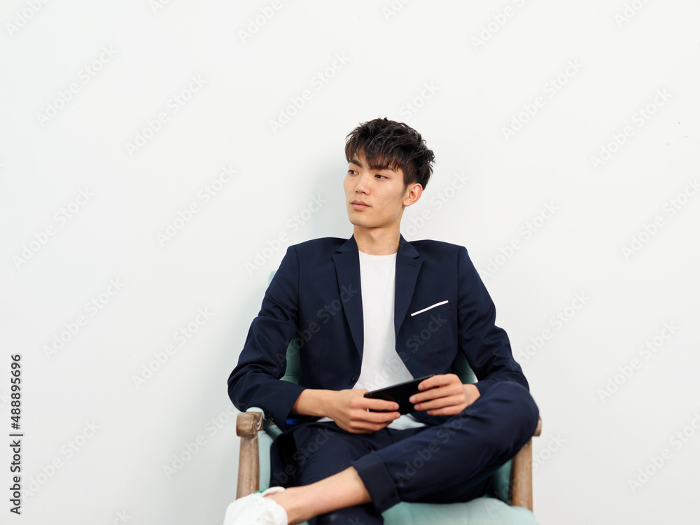 Portrait of handsome Chinese young man in dark blue leisure suit sitting in armchair and posing against white wall background. Legs crossed with mobile phone in hand, looking away and thinking. Portrait of handsome Chinese young man in dark blue leisure suit sitting in armchair and posing against white wall background. Legs crossed with mobile phone in hand, looking away and thinking.