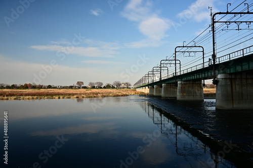 広がる冬の青空と浮かぶ白い雲、ゆったりと流れる多摩川、川を渡る鉄橋と川面に映る鉄橋の姿。 穏やかな流れの川面に写る青空と鉄橋が映り込む美しい風景写真。 彩度高め、色濃いめ、コピースペース広め、