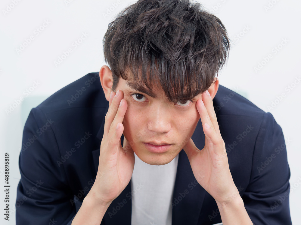 Portrait of handsome Chinese young man in dark blue leisure suit sitting in armchair and posing against white wall background. Two hands on his face and looking seriously, front view. Portrait of handsome Chinese young man in dark blue leisure suit sitting in armchair and posing against white wall background. Two hands on his face and looking seriously, front view.
