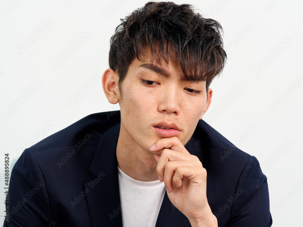 Portrait of handsome Chinese young man in dark blue leisure suit sitting in armchair and posing against white wall background. Looking away seriously with hand on his chin, front view. Portrait of handsome Chinese young man in dark blue leisure suit sitting in armchair and posing against white wall background. Looking away seriously with hand on his chin, front view.