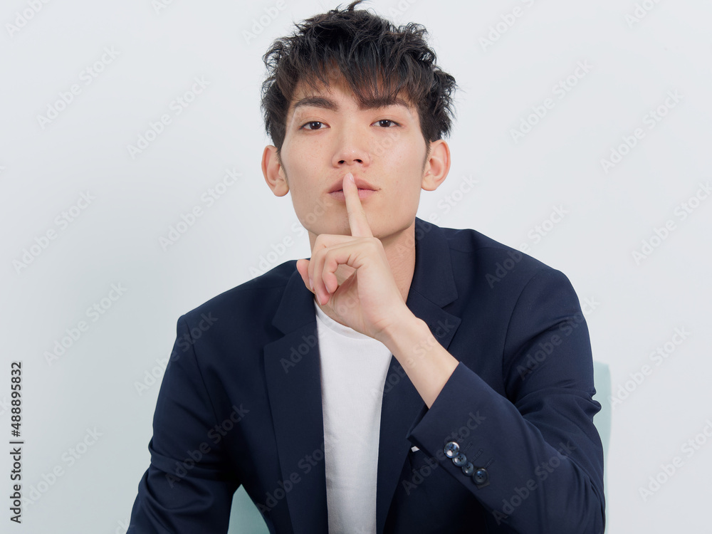 Portrait of handsome Chinese young man in dark blue leisure suit sitting in armchair and posing against white wall background. Finger on lips to make silent or be quiet gesture, front view. Portrait of handsome Chinese young man in dark blue leisure suit sitting in armchair and posing against white wall background. Finger on lips to make silent or be quiet gesture, front view.