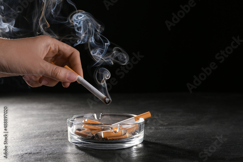 Obraz na plátně Woman holding smoldering cigarette over glass ashtray at grey table against blac