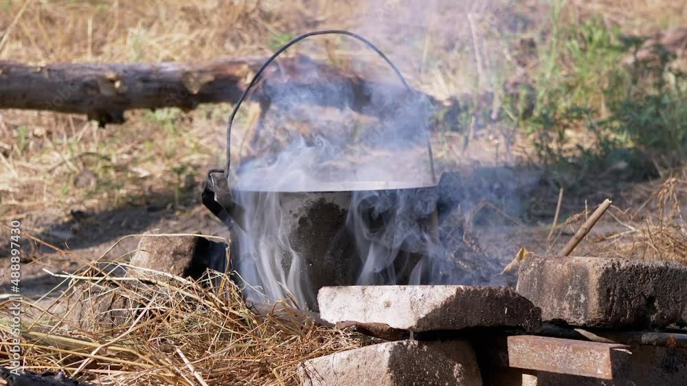 Cooking Food in a Saucepan Over an Open Bonfire in Smoke at Sunset. A black tourist boiler in soot smokes in sunlight on dry grass. Concept of camping, tourist recreation, nature survival. Summertime. Cooking Food in a Saucepan Over an Open Bonfire in Smoke at Sunset. A black tourist boiler in soot smokes in sunlight on dry grass. Concept of camping, tourist recreation, nature survival. Summertime.
