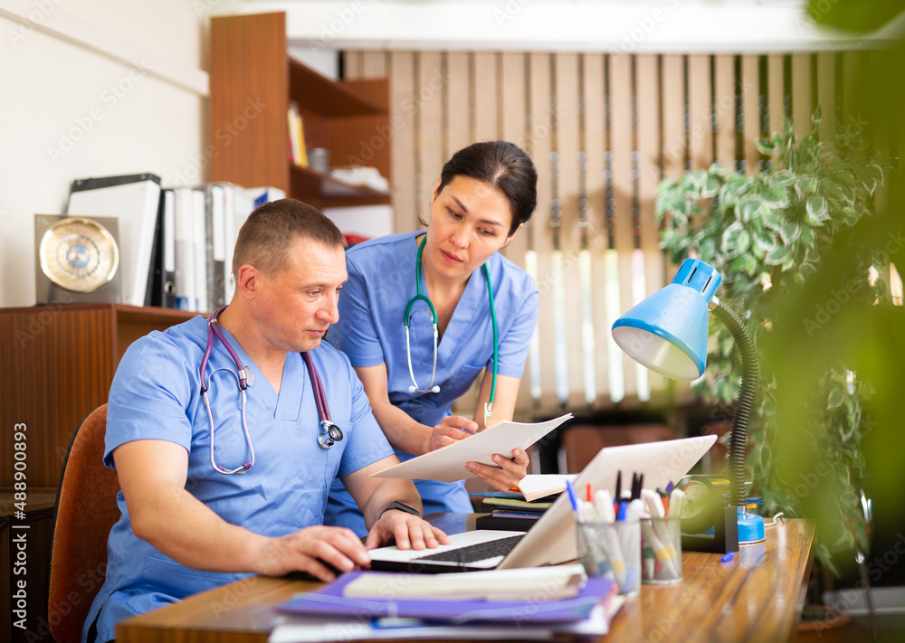 Obraz premium Portrait of two fellow doctors discussing clinical diagnosis of patients and filling up medical forms on laptop in office
