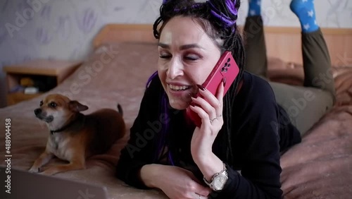The girl is talking using a mobile phone and smiling while lying on the bed.