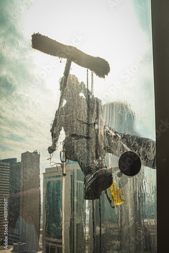 Skyscraper window cleaner hanging from a harness in Doha, Qatar. View from inside of the building behind a curtain of splashing water over the glass.  photo