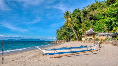 A traditional Filipino wooden outrigger boat called a banca sits on a beautiful tropical island sandy beach near resort buildings on the northern coast of Occidental Mindoro province, Philippines. photo