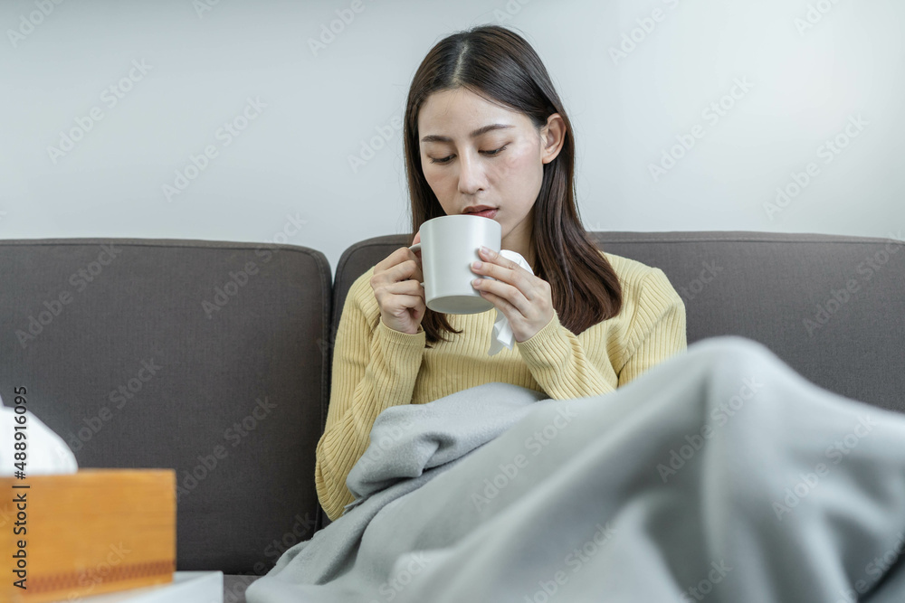 Sick, hurt or pain asian young woman, girl sore throat with glass, mug of warm water, headache have a fever, flu in weakness, sitting relaxed on sofa bed at home. Health care person on virus seasonal.