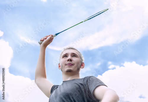 Portrait of a blond guy with a badminton racket in his hand on a pitch against cloudy sky