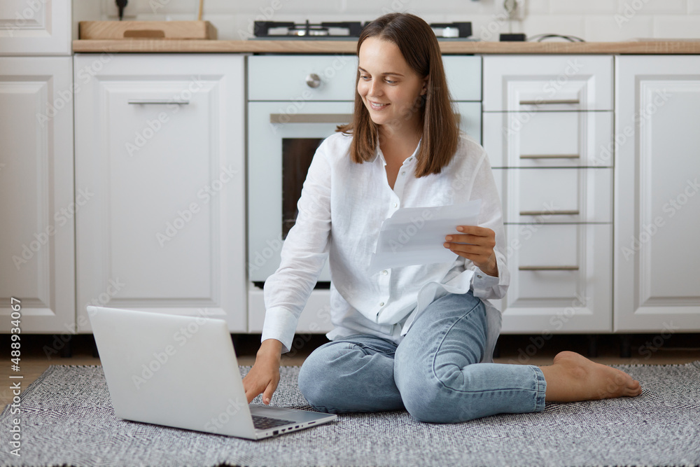 Portrait of smiling dark haired woman wearing white shirt and jeans sitting on floor, holding document in hand, looking at notebook screen with smile, working online at home. Portrait of smiling dark haired woman wearing white shirt and jeans sitting on floor, holding document in hand, looking at notebook screen with smile, working online at home.