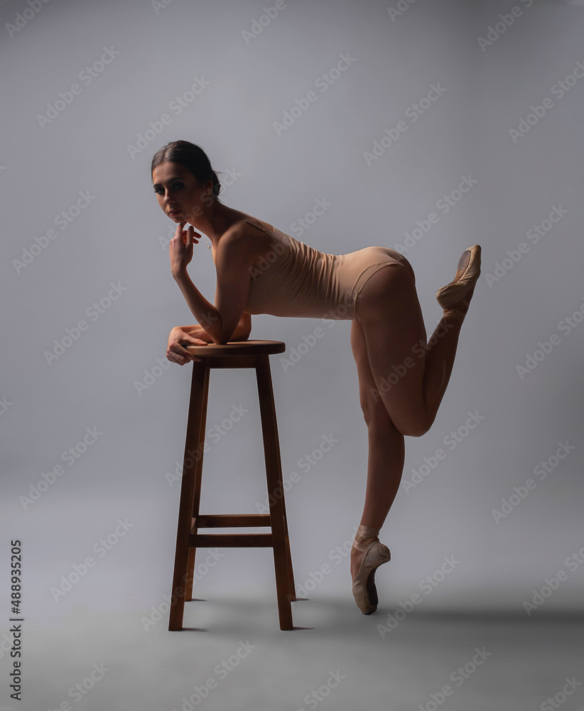 ballerina in a beige swimsuit near wooden chair on a gray background ballerina in a beige swimsuit near wooden chair on a gray background