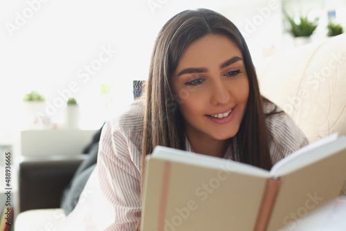 Young attractive woman reading book laying on couch at home, interesting story