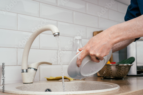 Close up of unrecognizable man in blue shirt washing up white round plate in beige marble kitchen sink under running water from faucet standing in white modern kitchen. Housework, cleaning concept.