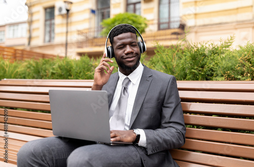 African american businessman in wireless headphones using laptop and listening music with closed eyes, sitting outdoors