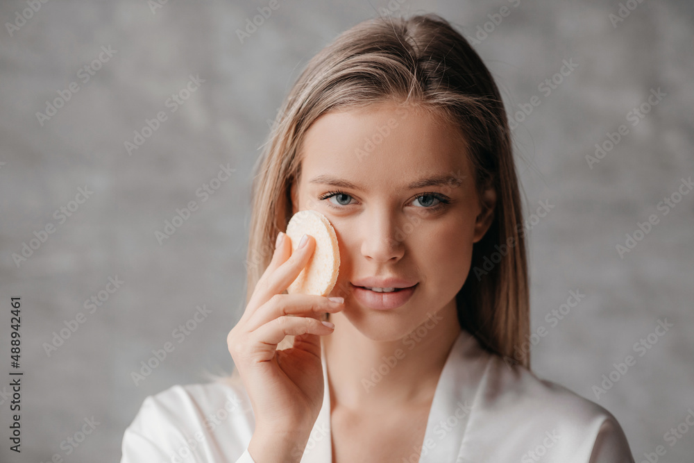 Skincare concept. Young woman using cosmetic sponge while doing facial skincare routine, standing in bathroom Skincare concept. Young woman using cosmetic sponge while doing facial skincare routine, standing in bathroom