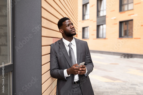 Strategic thinking. Thoughtful african american businessman holding smartphone and looking away, leaning on wall