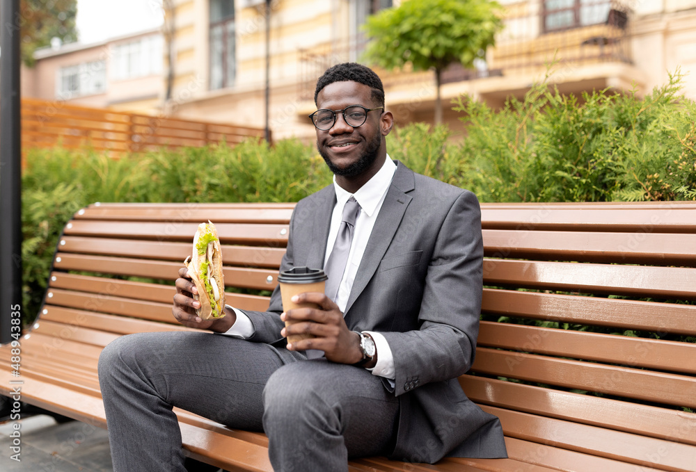 Fototapeta premium Enjoy your lunch. Happy african american businessman drinking coffee and eating sandwich, sitting on bench