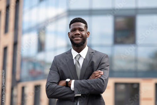 Wealthy african american entrepreneur young man standing outside modern office building, posing with folded arms