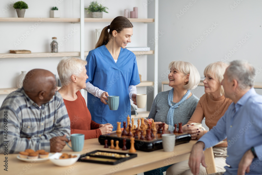 Friendly nurse holding mugs, talking to elderly people, serving tea