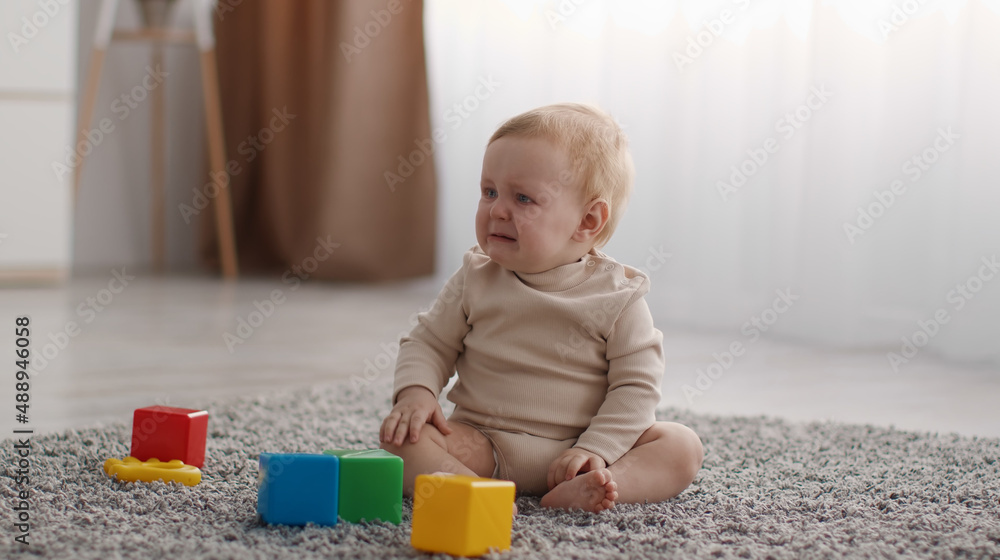 Portrait of cute little baby crying, sitting on floor with colorful cubes, suffering from pain or loneliness, free space Portrait of cute little baby crying, sitting on floor with colorful cubes, suffering from pain or loneliness, free space