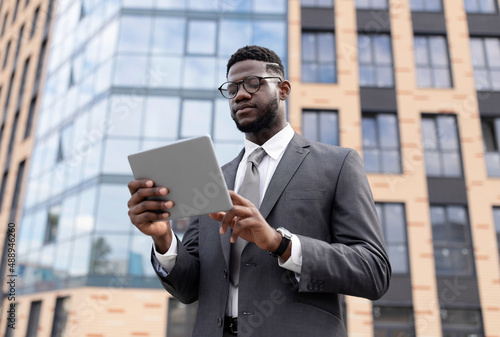 Focused black man in formal wear standing by office building, holding digital tablet, using business application