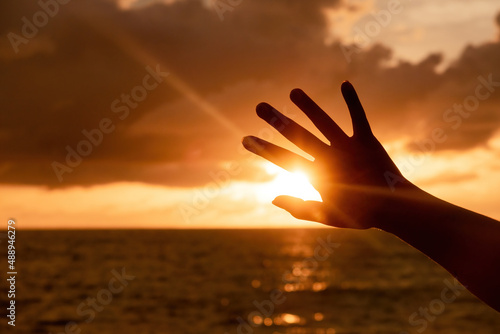 Hand raised in silhouette shadow block golden sun, main scene is sky and sea.