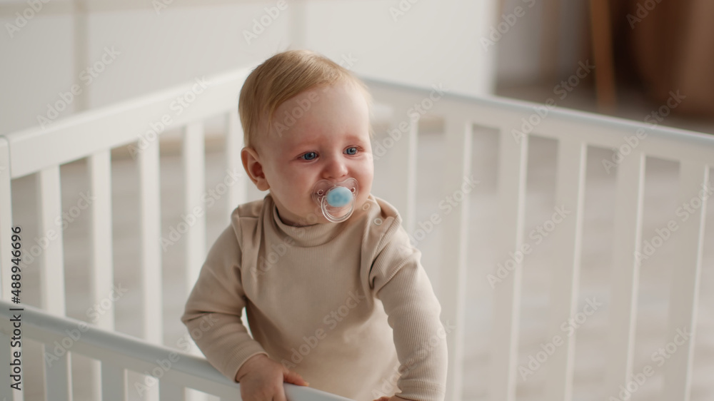 Sedative for toddlers. Little baby with pacifier standing in crib, looking aside at bedroom, panorama, free space