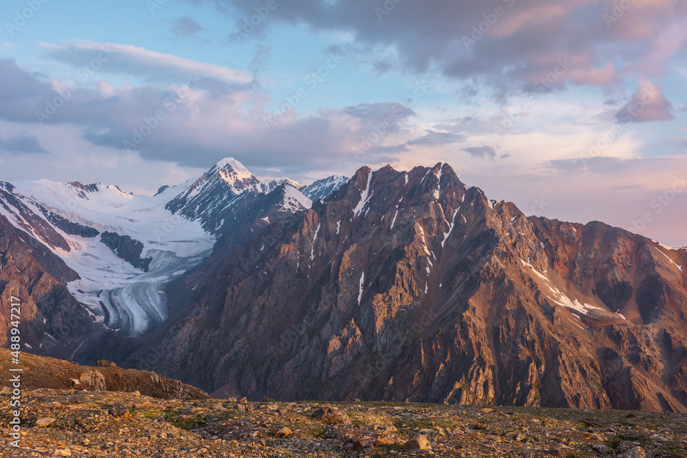 Naklejka premium Vivid alpine landscape with high snow mountains and large glacier in sunrise colors. Colorful mountain scenery with sunlit golden rocks and mountains at sunrise. Early morning at very high altitude.