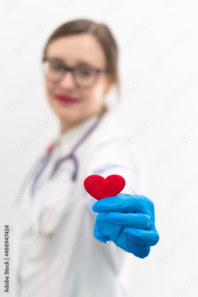 Young pretty woman doctor in a white medical coat and rubber gloves with a phonendoscope holds a red heart in her hands in a hospital. Selective focus. Portrait