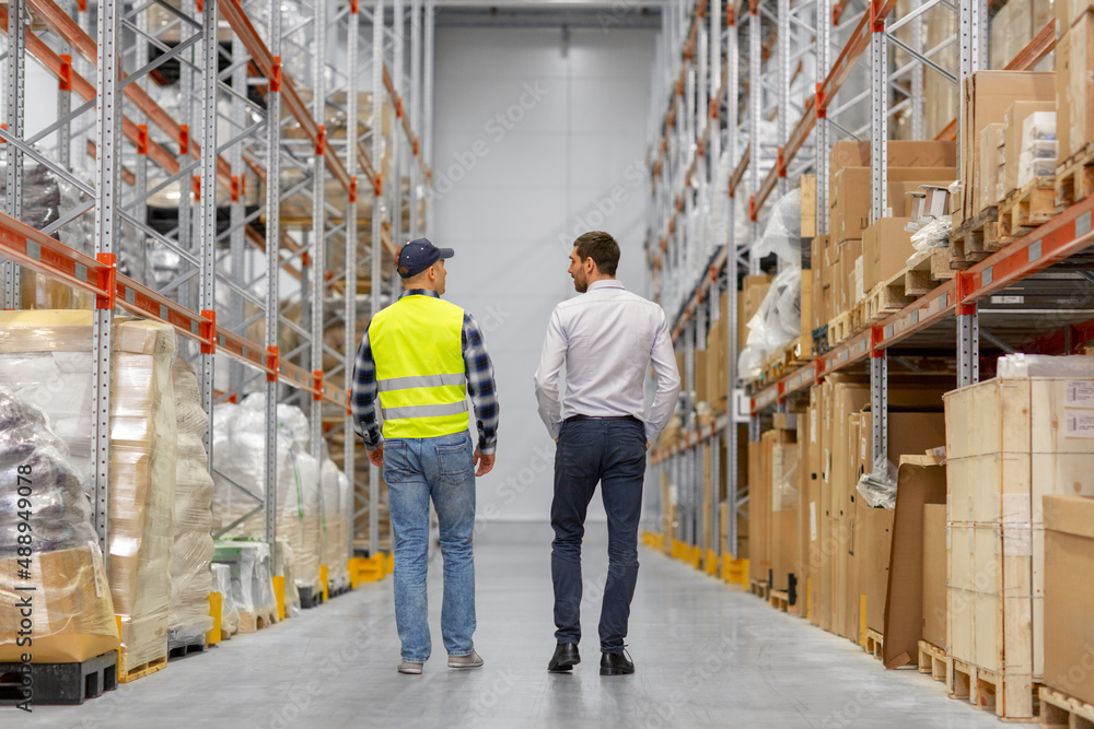 wholesale, logistic business and people concept - manual worker and businessman walking along warehouse wholesale, logistic business and people concept - manual worker and businessman walking along warehouse