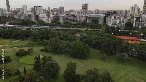 Train departing from station close to golf and tennis court with buildings in background, Buenos Aires. Aerial forward photo