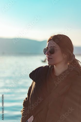 Cute girl posing in front of sea. She getting cold. Winter season. Vertical photo. Sunset at sea. Her hair blowing with wind. She has sun glasses.