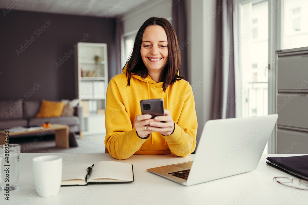 Smiling female freelancer using smart phone while sitting with laptop at home office Smiling female freelancer using smart phone while sitting with laptop at home office
