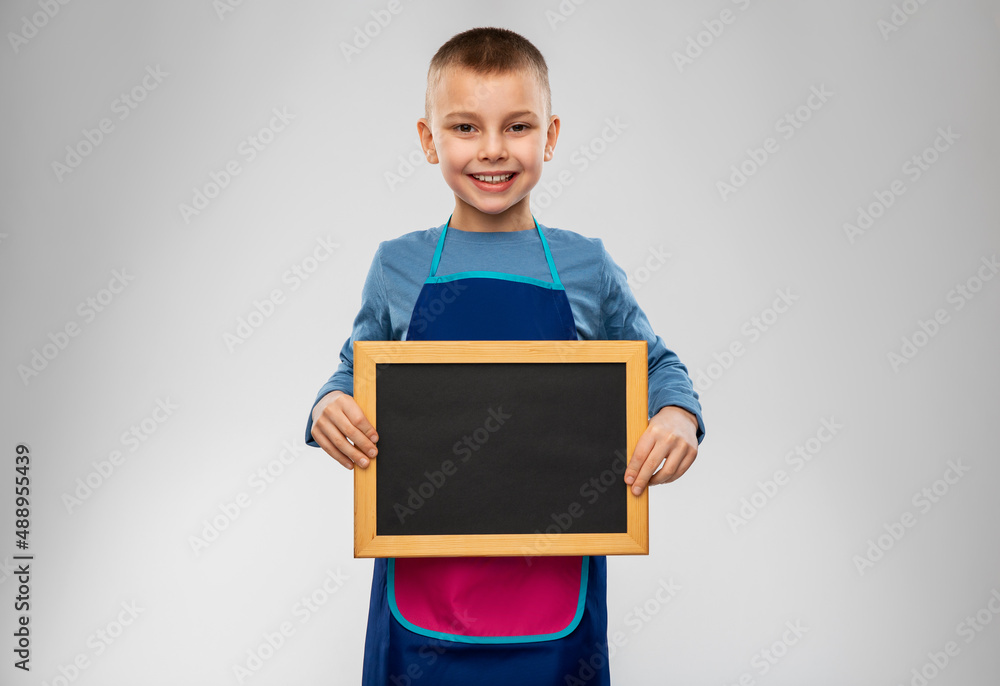 cooking, culinary and profession concept - happy smiling little boy in apron holding chalkboard over grey background cooking, culinary and profession concept - happy smiling little boy in apron holding chalkboard over grey background