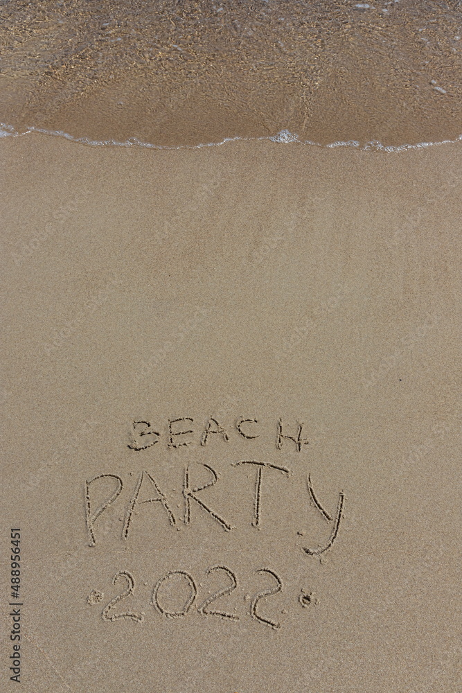 Fototapeta premium The inscription beach party 2022 on the sand by the water and the rising wave, seashore beach vacation by the sea