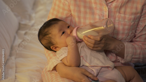 Photos close up innocent hungry infant covered with burping cloth is lying in her mother’s arms and drinking milk from bottle in the bedroom