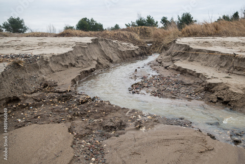 Stream of rainwater broke through sandy barrier and hillock with dry grass, flowing among sand, pebbles, rubble and shells along winding riverbed on background of meadow with trees and cloudy sky.