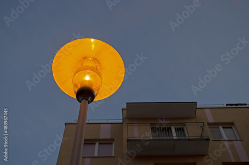Round-shaped lantern on a black holder was lit on background of blue twilight sky and upper floors of apartment building with a yellow facade early in the morning at dusk. Photo taken at blue hour.