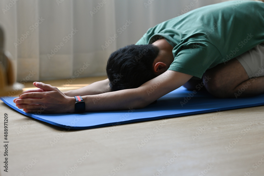 A side view portrait of a young man stretching on a mat in a living room, for home exercise, yoga and a class concept.