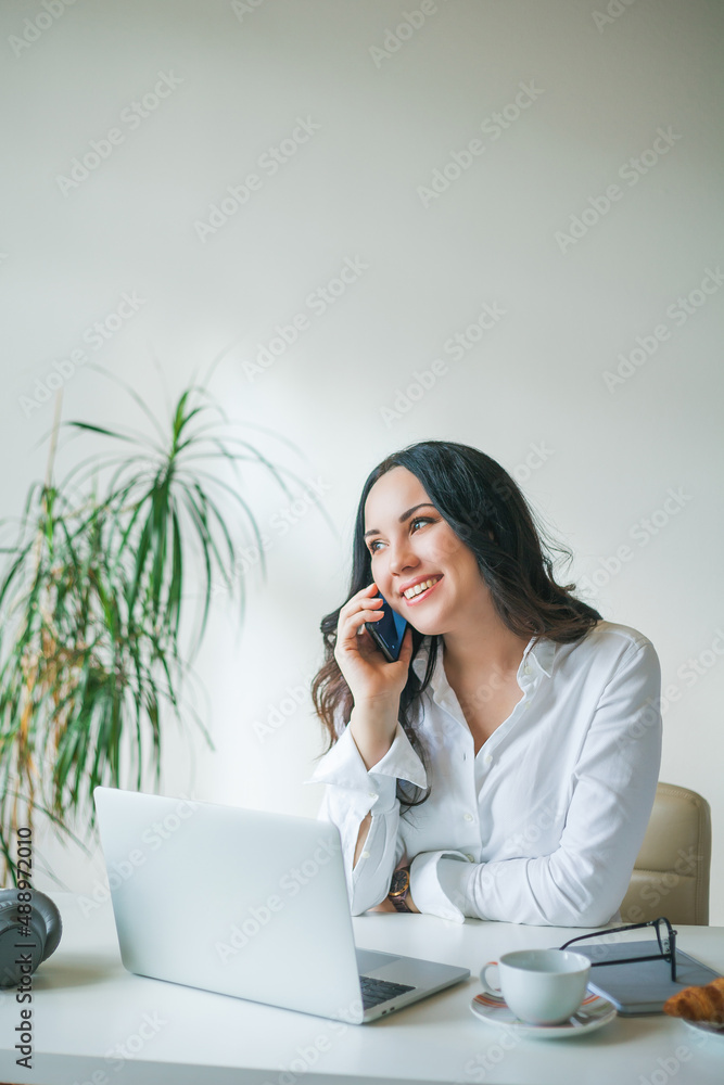 Young businesswoman works at a computer and speaks on the phone. A beautiful dark-haired woman in a white shirt works online in a bright office. Drinking coffee and eating a croissant. Young businesswoman works at a computer and speaks on the phone. A beautiful dark-haired woman in a white shirt works online in a bright office. Drinking coffee and eating a croissant.