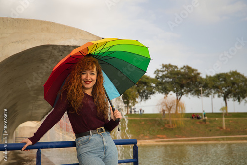 Portrait of young woman, red hair, freckles, with a rainbow umbrella, under a waterfall, in an outdoor park. Concept color, happiness, well-being, fun, rain.