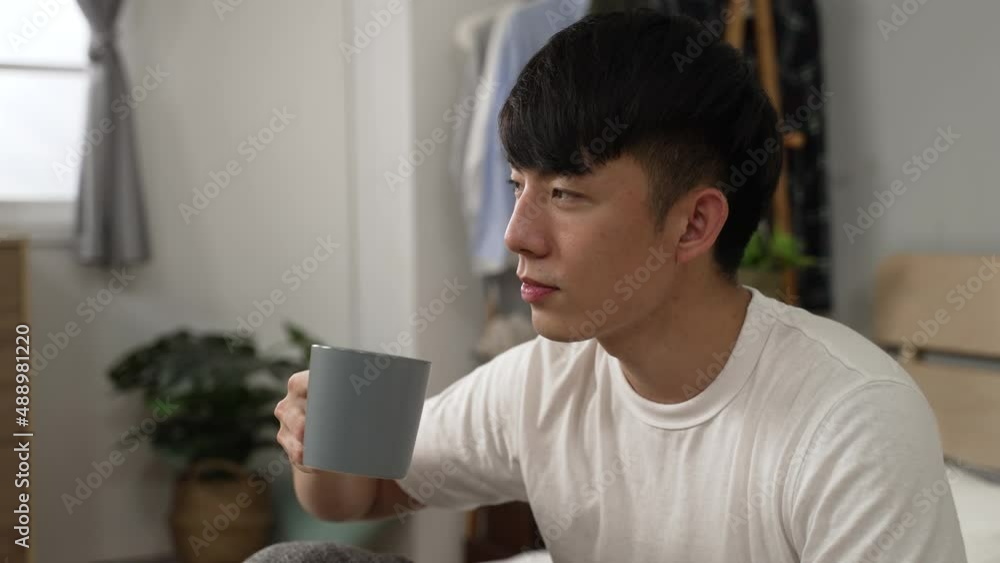 headshot of a happy asian young man enjoying morning tea and looking into distance with a smile after waking up in the bedroom at home headshot of a happy asian young man enjoying morning tea and looking into distance with a smile after waking up in the bedroom at home