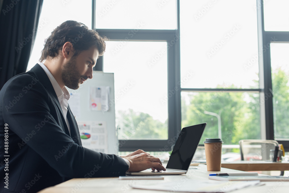 side view of bearded businessman typing on laptop with blank screen in office side view of bearded businessman typing on laptop with blank screen in office